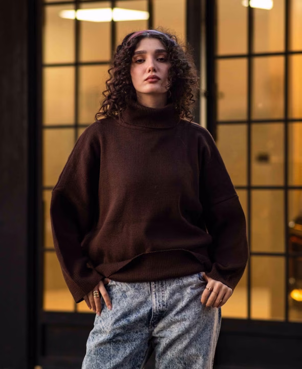 Woman with curly hair wearing a brown turtleneck sweater and light blue jeans standing in front of a glass door.