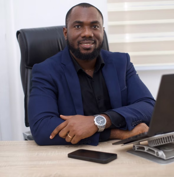 Smiling man in a dark blue blazer sitting at a desk with a laptop and smartphone.