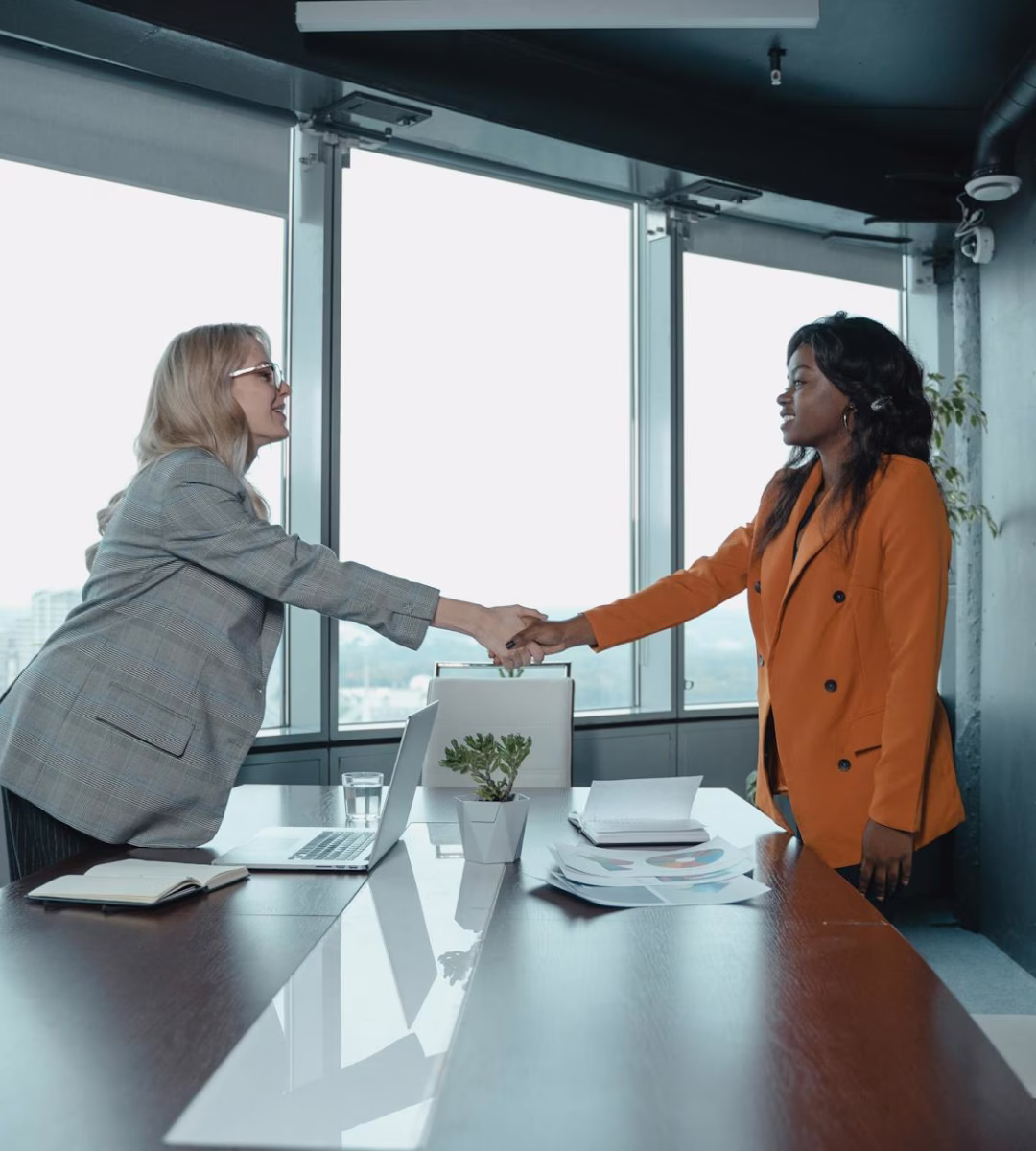 Two professional women shaking hands across a conference table in a modern office with large windows.