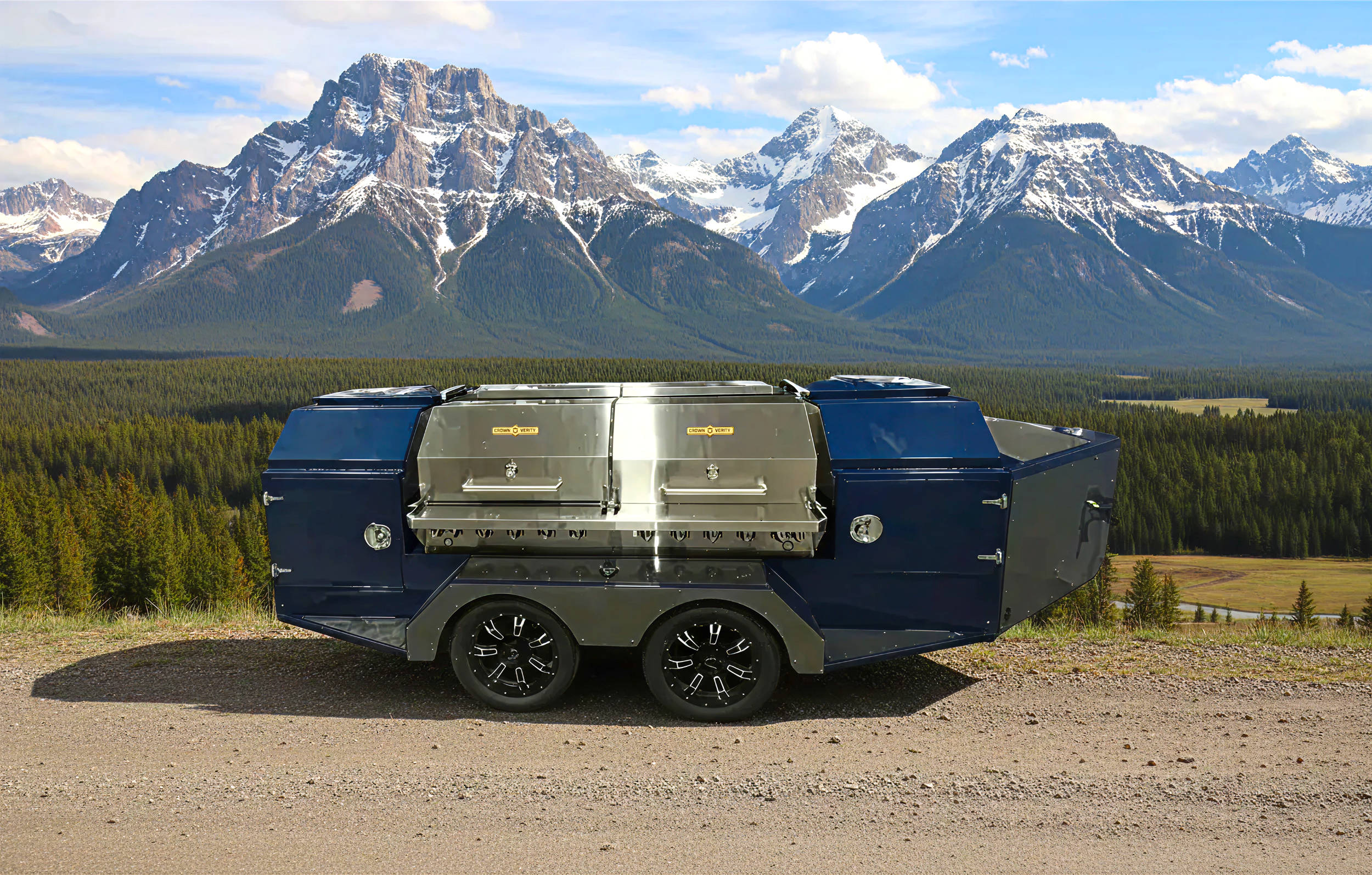 Custom blue and stainless steel BBQ trailer with dual Crown Verity grills parked on a gravel road with snow-capped mountains in the background.