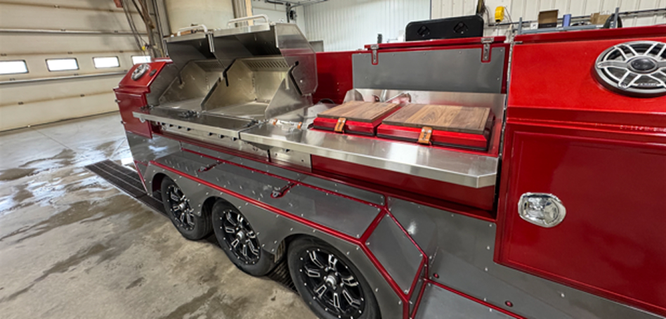 Open blue custom BBQ trailer with metal drawers and grill, set against a mountainous landscape with clear skies.