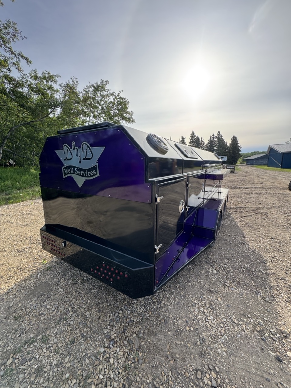 Blue custom barbecue (BBQ) trailer with double axles parked on a dirt path in a dense forest.