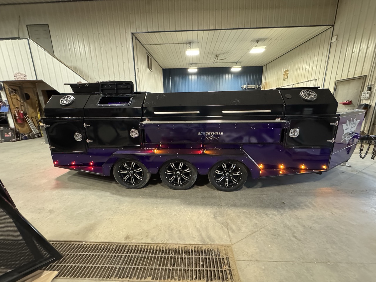 Blue custom barbecue (BBQ) trailer with double axles parked on a dirt path in a dense forest.