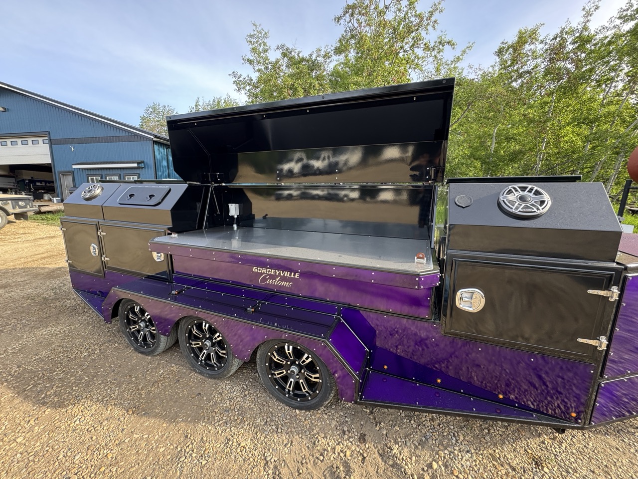 Blue custom barbecue (BBQ) trailer with double axles parked on a dirt path in a dense forest.
