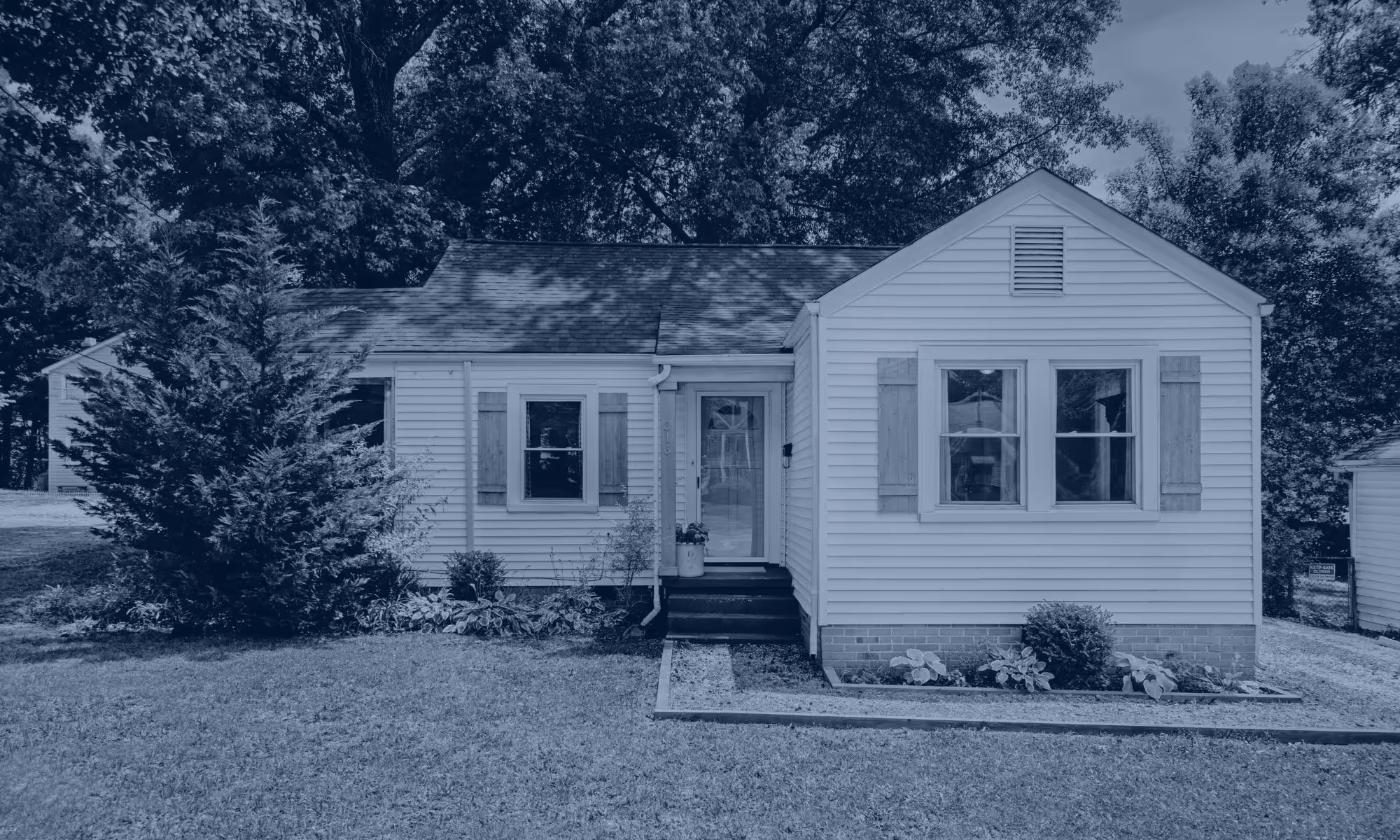 Single-story white house with a front porch, two windows with shutters, and surrounded by trees and a grassy yard.