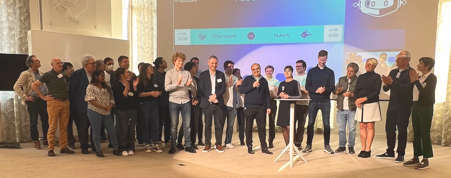 A group of around 20 people standing on a stage, with two individuals holding microphones at a podium, in front of a large screen displaying a game score.