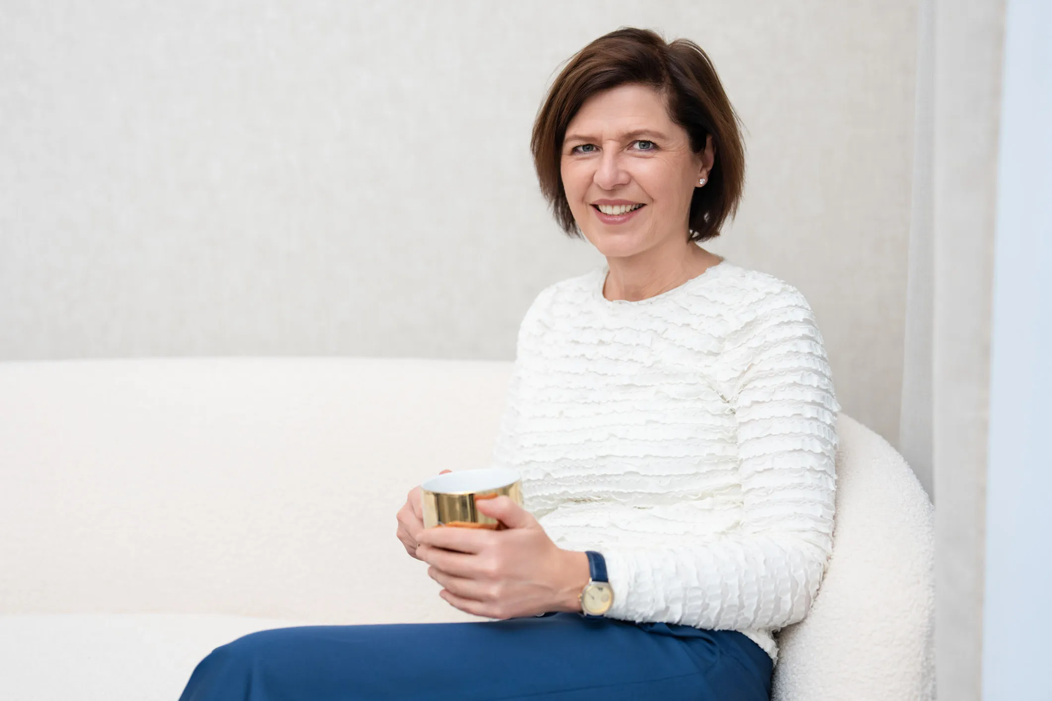 Woman in white textured top and blue pants sitting on a white couch holding a gold-rimmed cup.