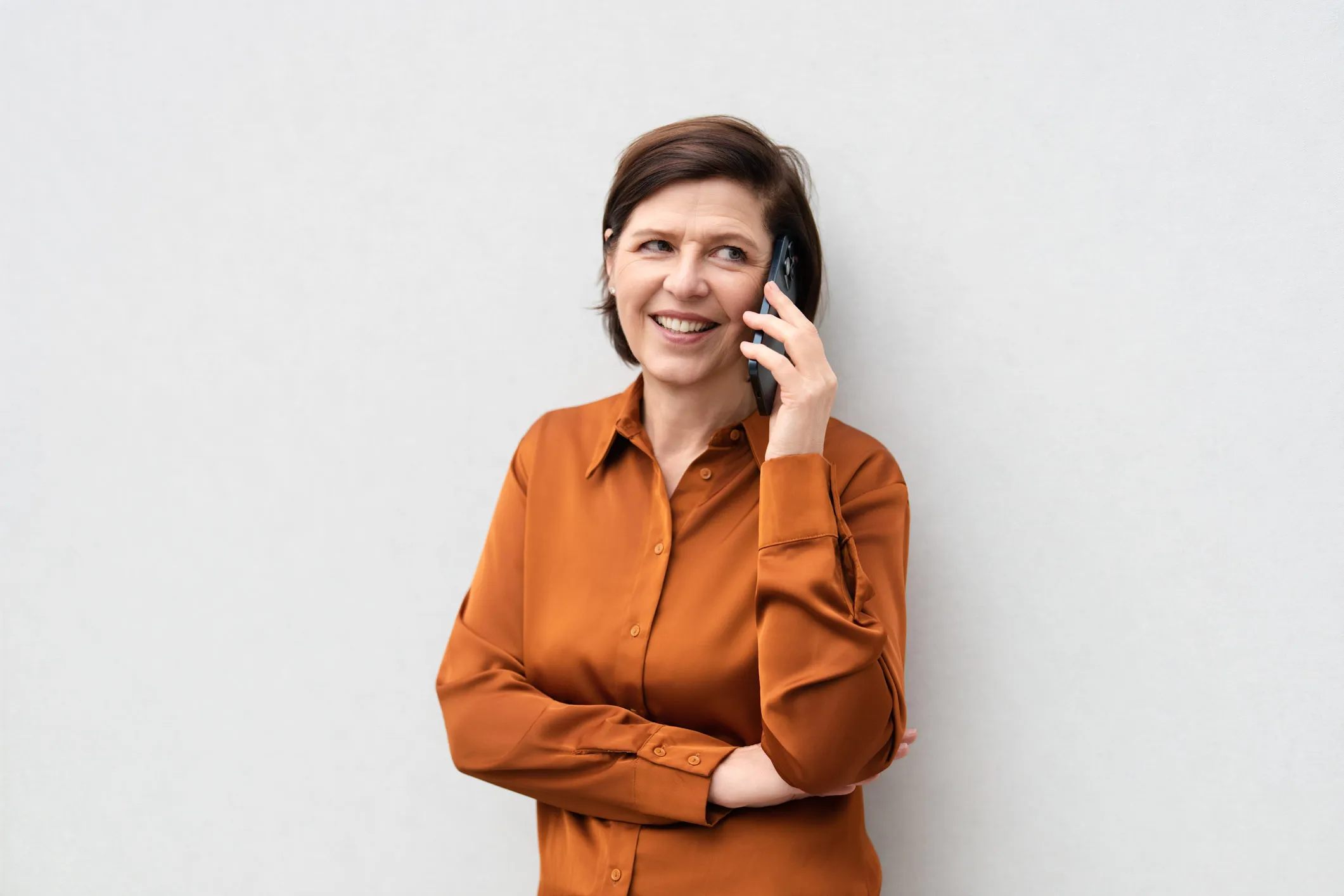 Smiling woman in an orange shirt talking on a smartphone against a plain light background.
