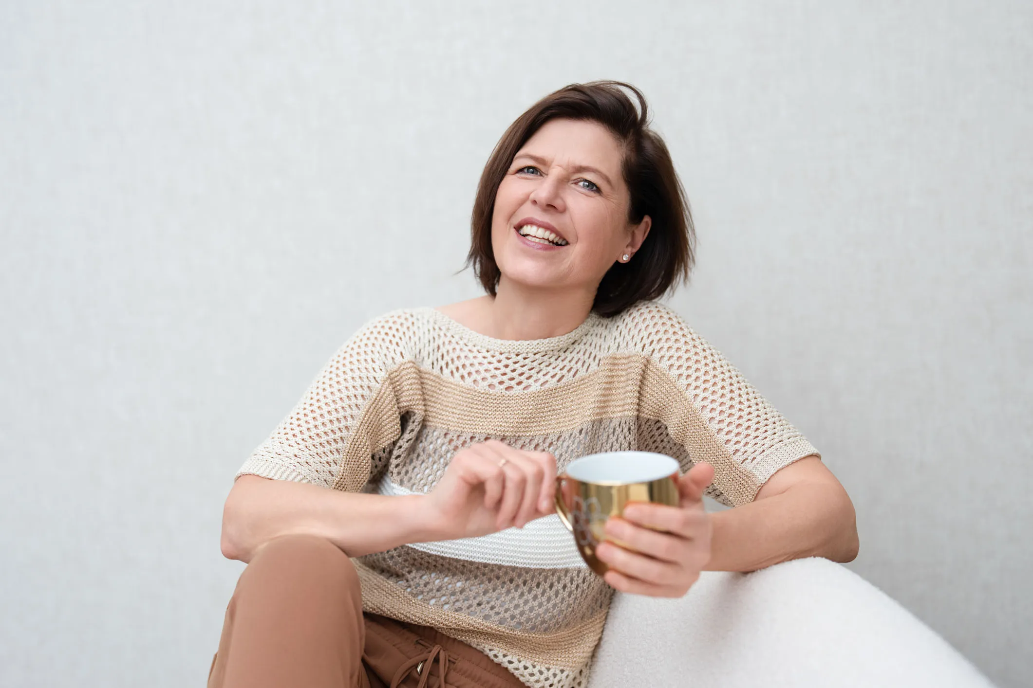 Smiling woman with short brown hair sitting on a white chair, holding a gold cup and wearing a beige and white knit top with brown pants.