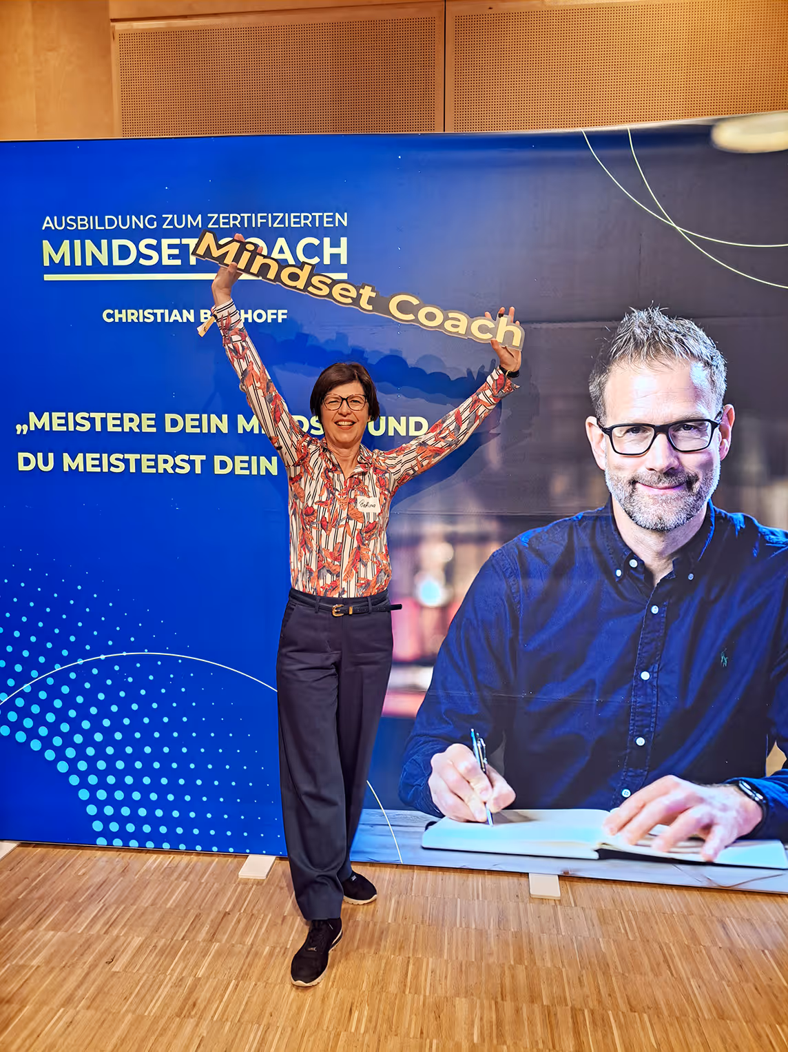 Woman standing with arms raised holding a 'Mindset Coach' sign in front of a blue backdrop advertising certified mindset coach training.