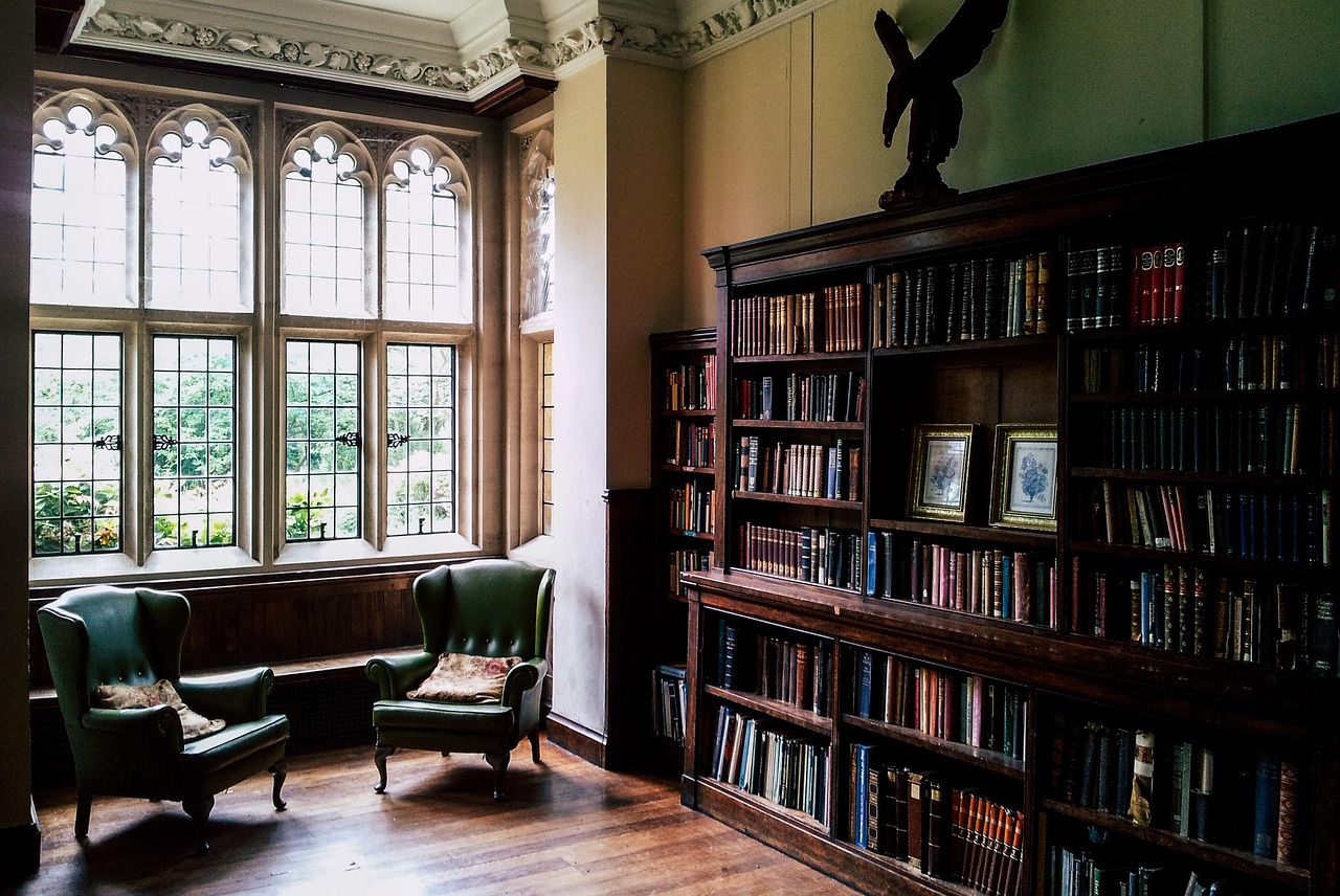 Cozy library corner with two green armchairs, large decorative window, and tall wooden bookshelf filled with books.