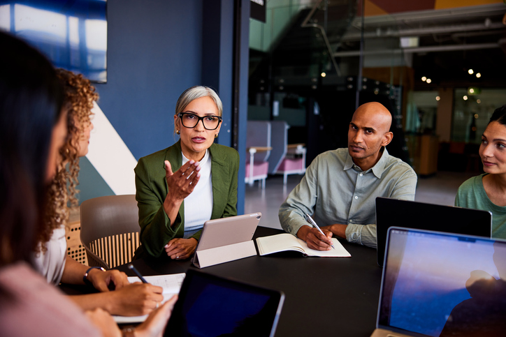 A diverse group of five professionals engaged in a discussion around a conference table with laptops and notebooks.