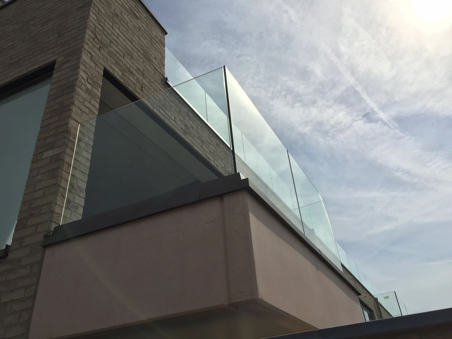 Glass railing on a balcony attached to a modern brick and stucco building under a partly cloudy sky.