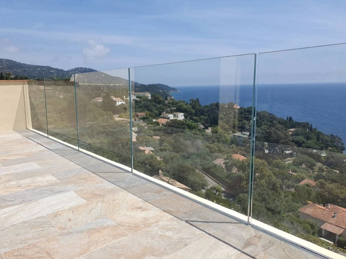 Balcony with beige tiled floor and clear glass railing overlooking a green coastal hillside and calm blue sea under a clear sky.