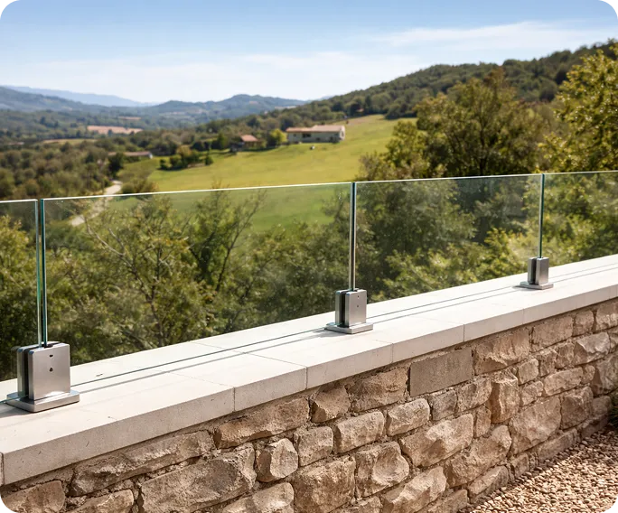 Glass railing with metal supports on stone wall overlooking green hills and countryside under a clear sky.