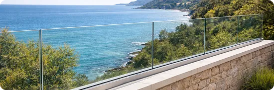 Glass railing overlooking a coastal view with blue ocean and green trees along the shoreline.