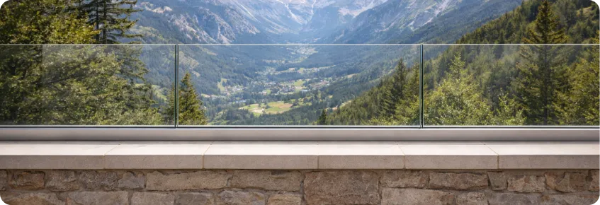 Stone terrace with glass railing overlooking a green valley surrounded by forested mountains under a blue sky.