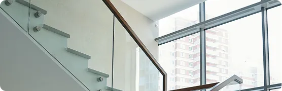 Modern staircase with glass railing and wooden handrail beside large windows with city view.