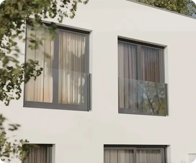 Modern white building facade with four large windows featuring sheer curtains and glass balcony railings, partially obscured by tree branches.