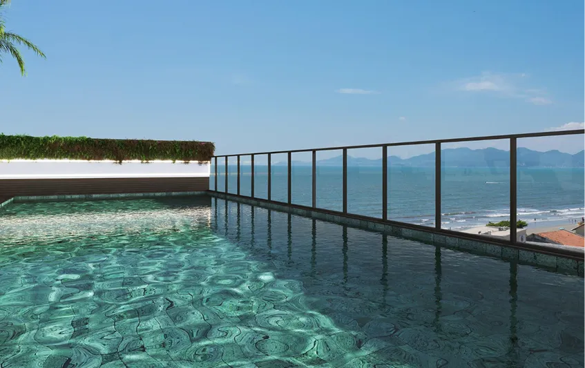 Infinity pool with clear turquoise water overlooking a beach and ocean, with glass railing and mountains in the distance under a blue sky.