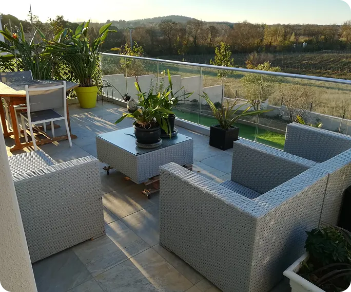 Outdoor balcony seating area with gray wicker chairs and table, potted plants, and glass railing overlooking a sunlit rural landscape.