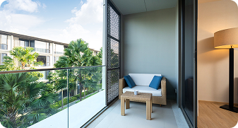 Modern balcony with a wooden chair, blue cushions, glass railing, and view of green palm trees and neighboring buildings.