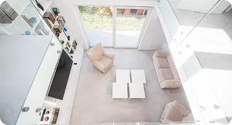 Bright modern living room with beige sofa, armchair, coffee tables, and built-in shelves under large windows.
