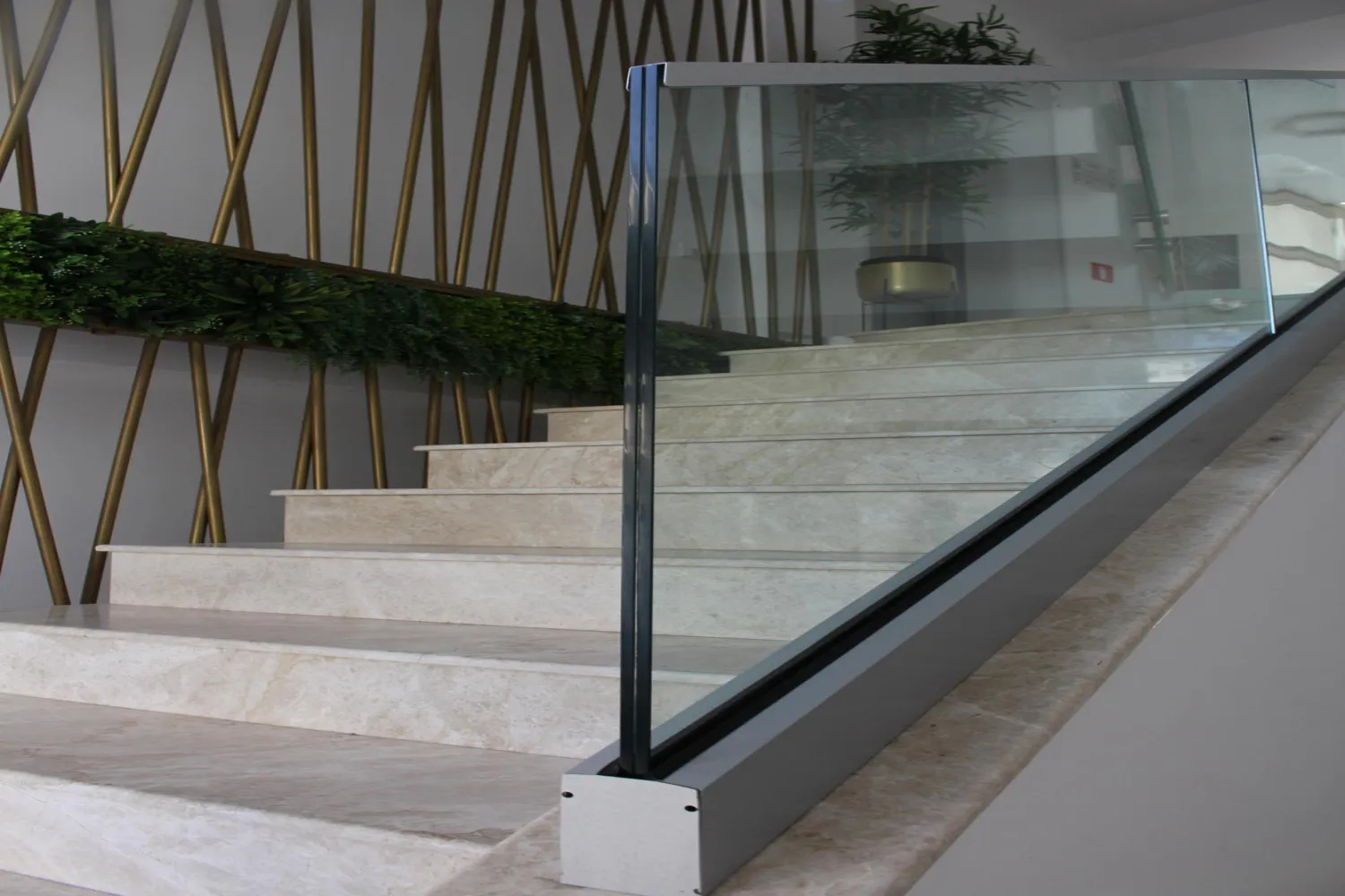 Modern indoor staircase with beige marble steps and glass railing, decorated with green plants and gold accents.