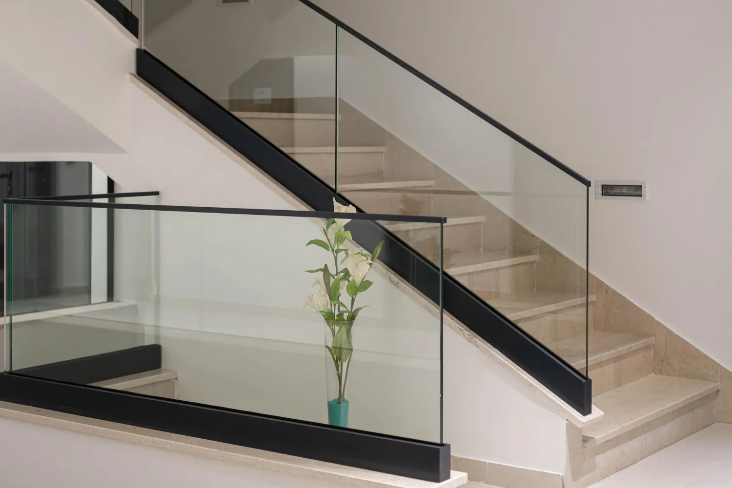 Indoor staircase with beige steps and glass railing featuring black metal supports and a vase with white flowers on the landing.