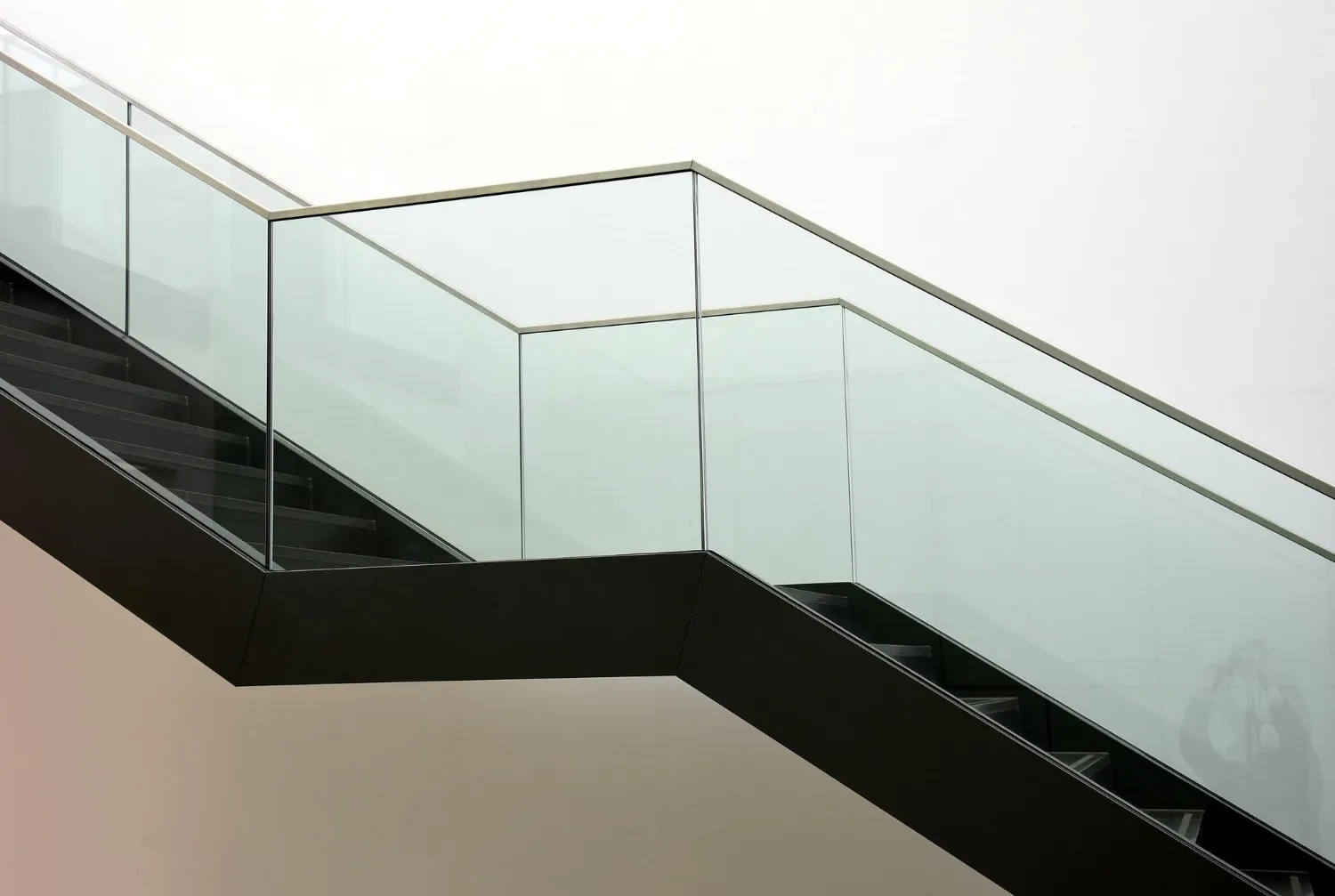 Modern staircase with black steps and transparent glass railing against a plain light background.