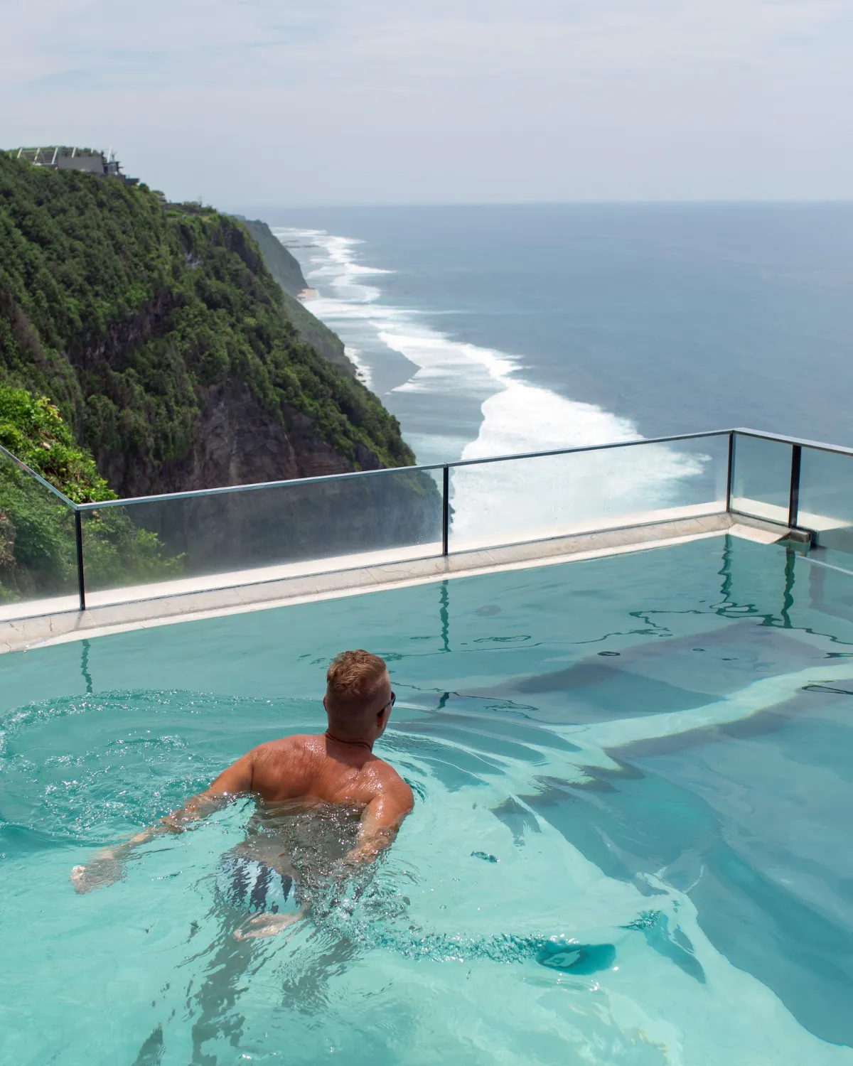 Man swimming in an infinity pool overlooking a green cliff and ocean coastline.
