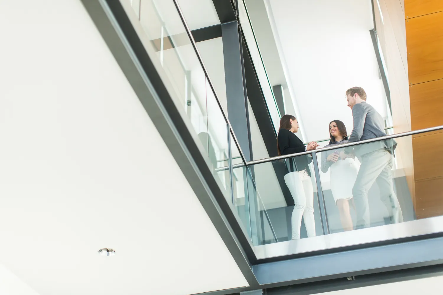 Three business professionals standing on a glass balcony having a conversation in a modern office building.