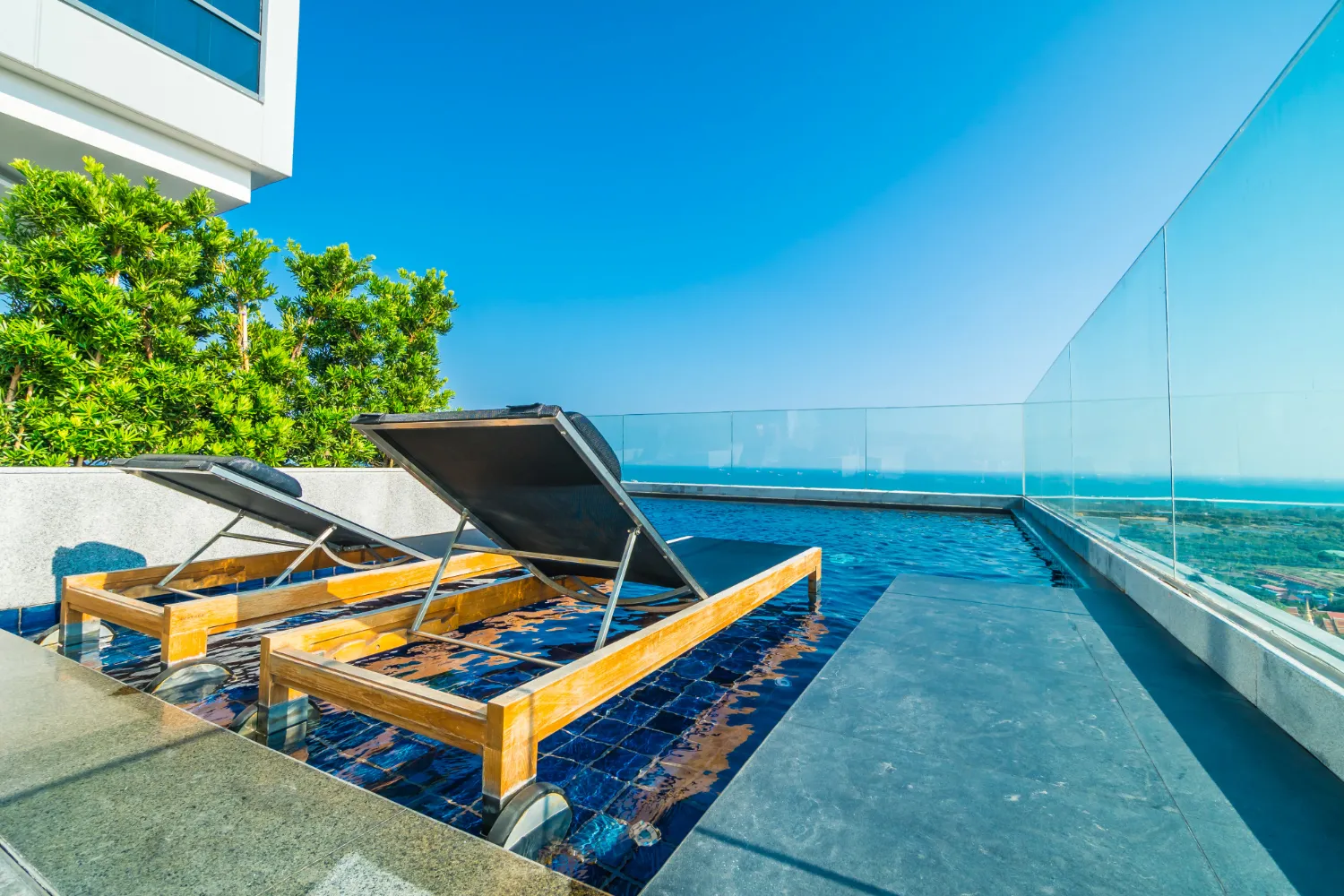 Two wooden sun loungers with black cushions partially submerged in a rooftop pool with glass railing and a clear blue sky.