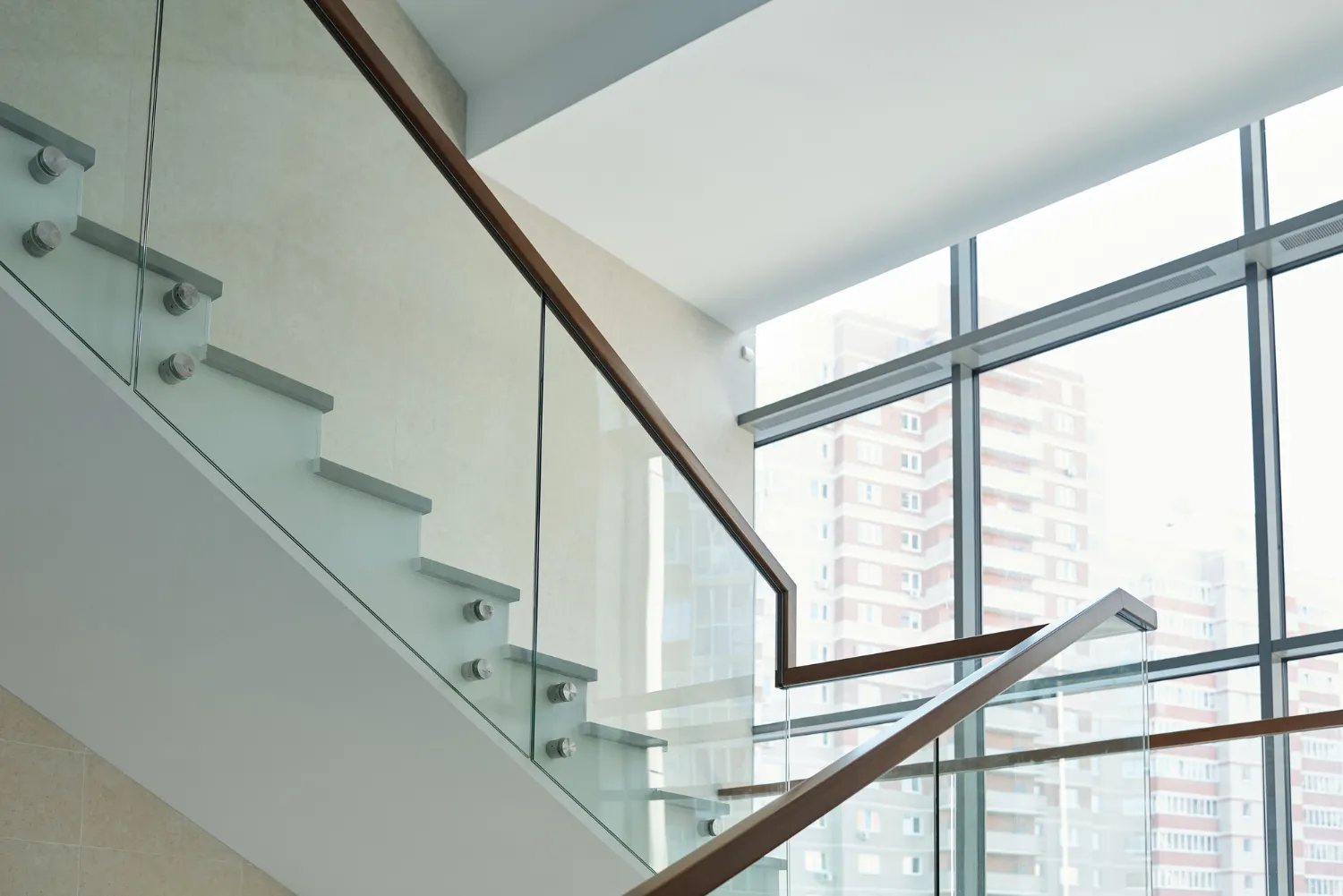 Modern indoor staircase with glass panels and wooden handrails next to large windows showing an urban building outside.
