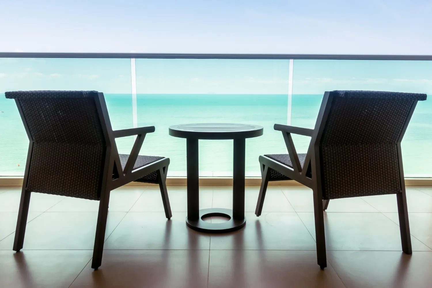 Two dark wicker chairs and a round table on a balcony overlooking a calm turquoise sea.