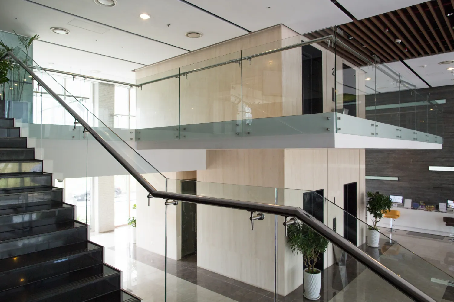 Modern office lobby with black staircase, glass railings, potted plants, and elevator doors on the second floor.