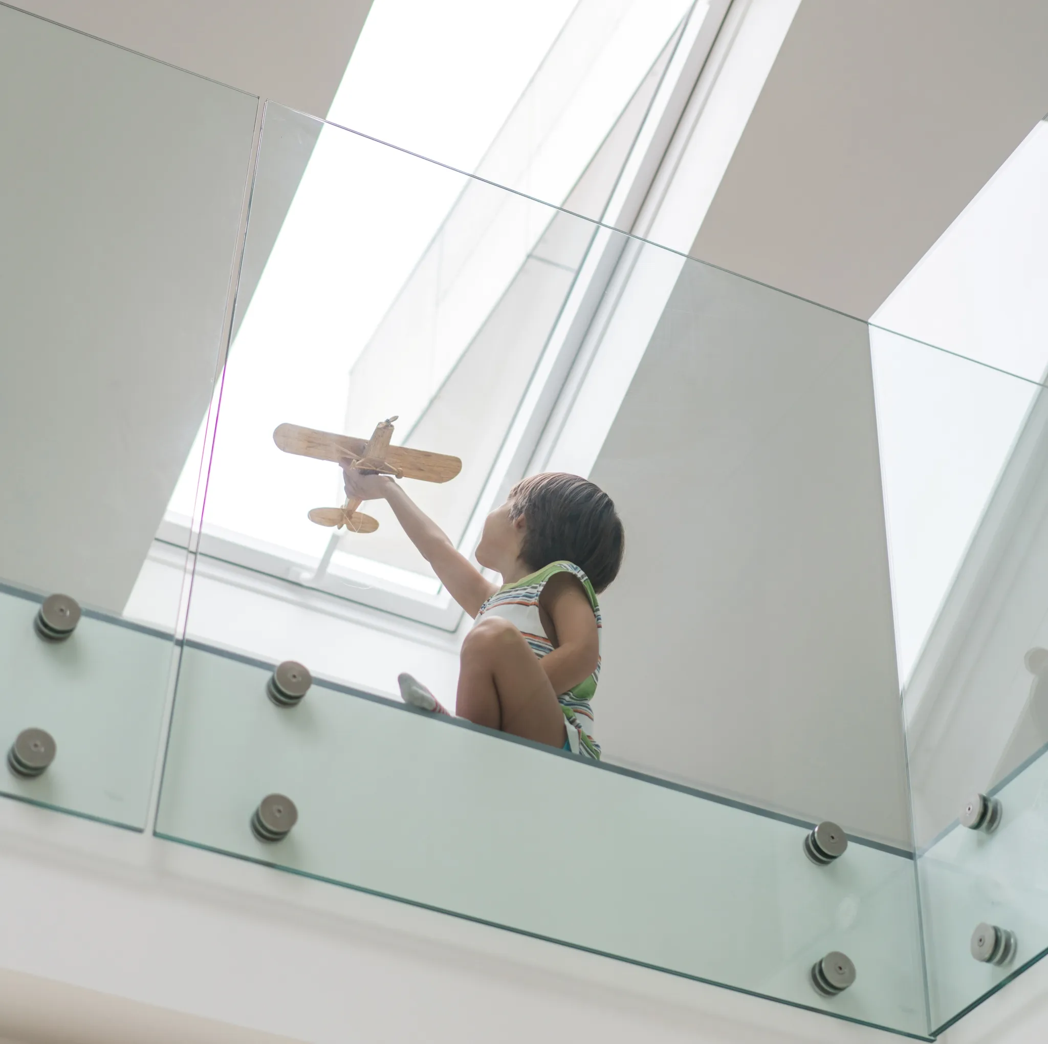 Child sitting behind a glass railing holding a wooden toy airplane up towards a bright skylight.