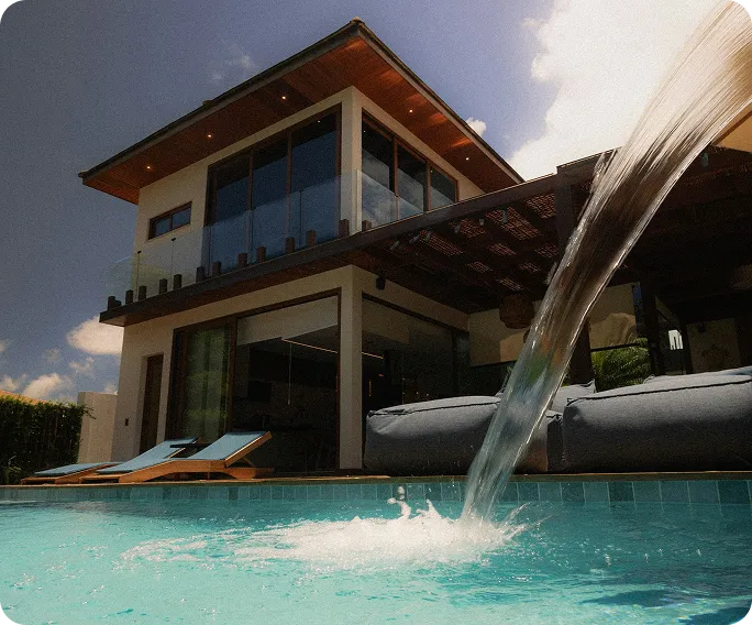 Modern two-story house with large glass windows, outdoor lounge chairs, and water flowing into a turquoise swimming pool.