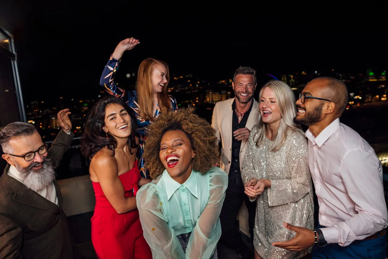 Group of seven diverse friends laughing and dancing at a nighttime rooftop party with city lights in the background.