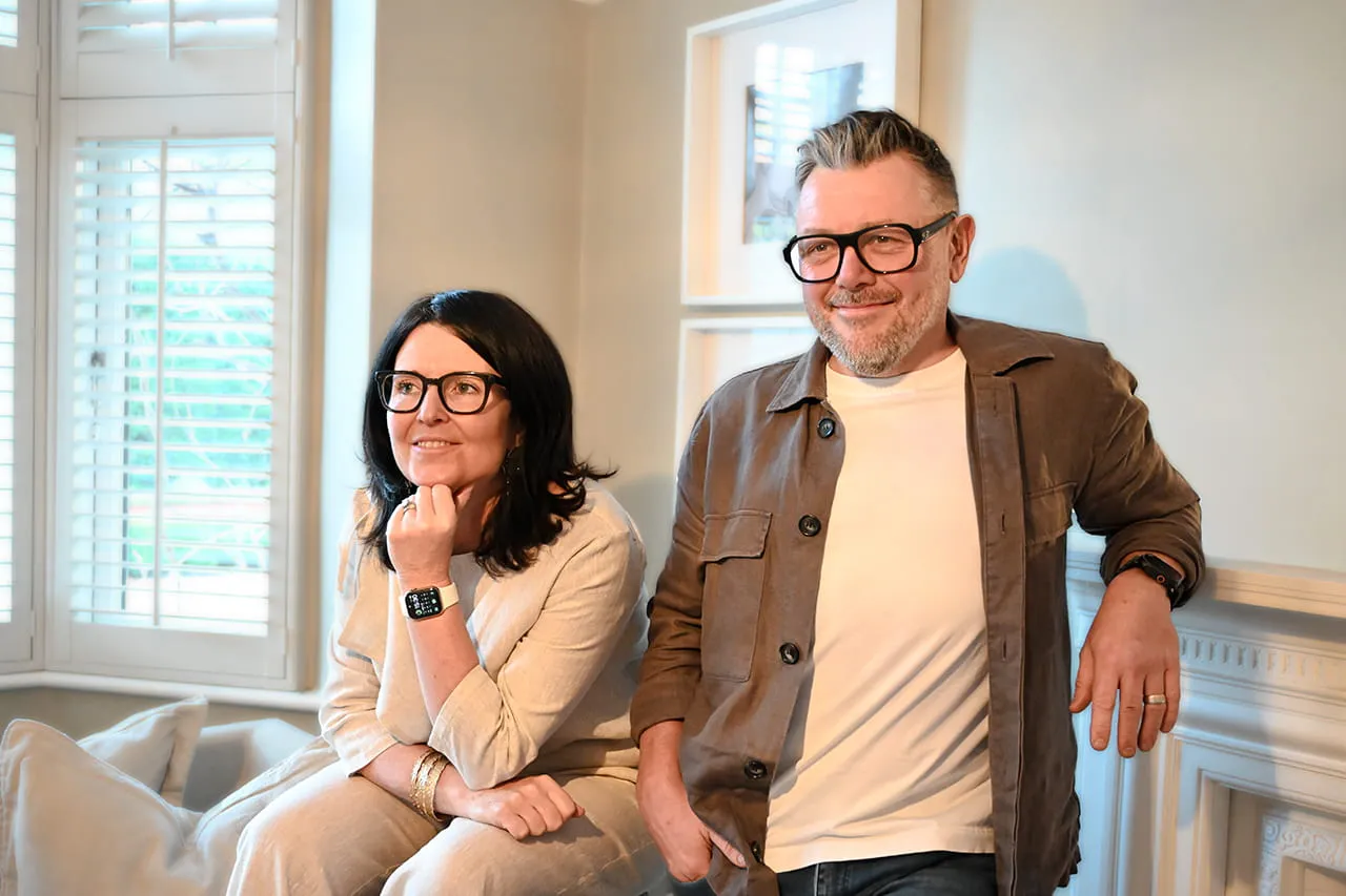 A woman and a man wearing glasses smiling indoors, with the woman sitting on a couch and the man leaning against a wall.