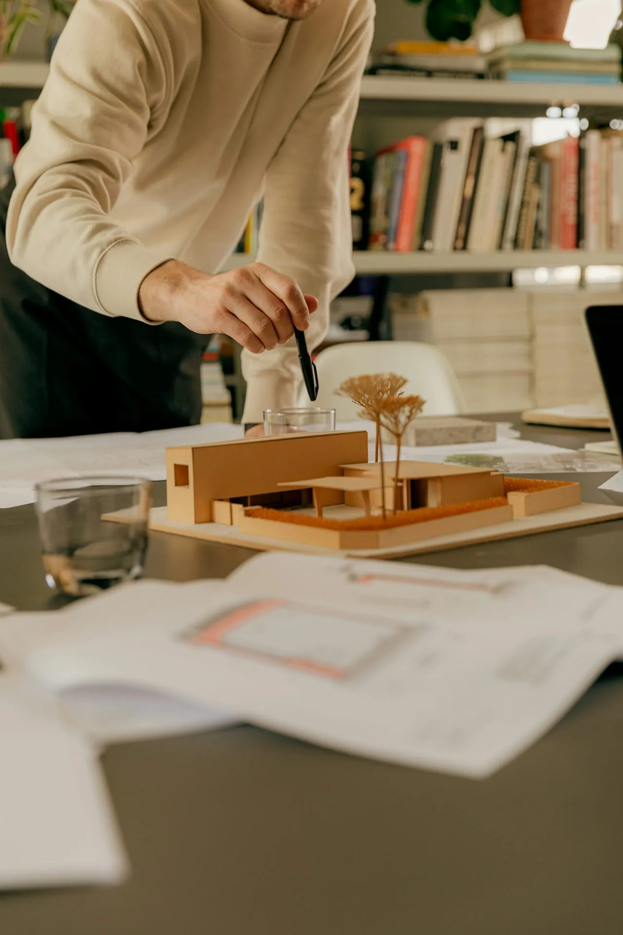 Person in beige sweater pointing at a wooden architectural model on a table with papers and books in the background.