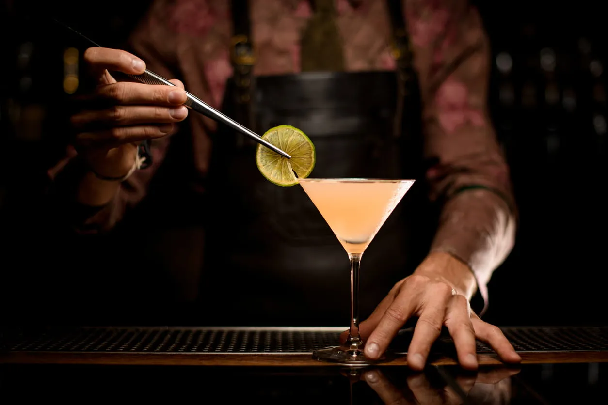 Bartender placing a lime wheel garnish on a pale pink cocktail in a martini glass.