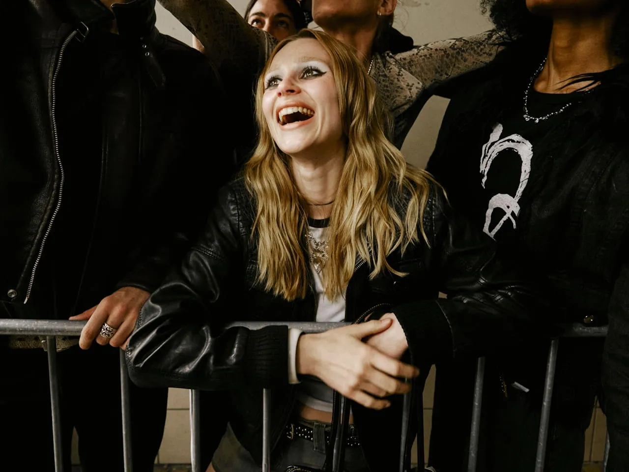 Smiling young woman with blonde hair leaning on a metal barricade surrounded by other people.
