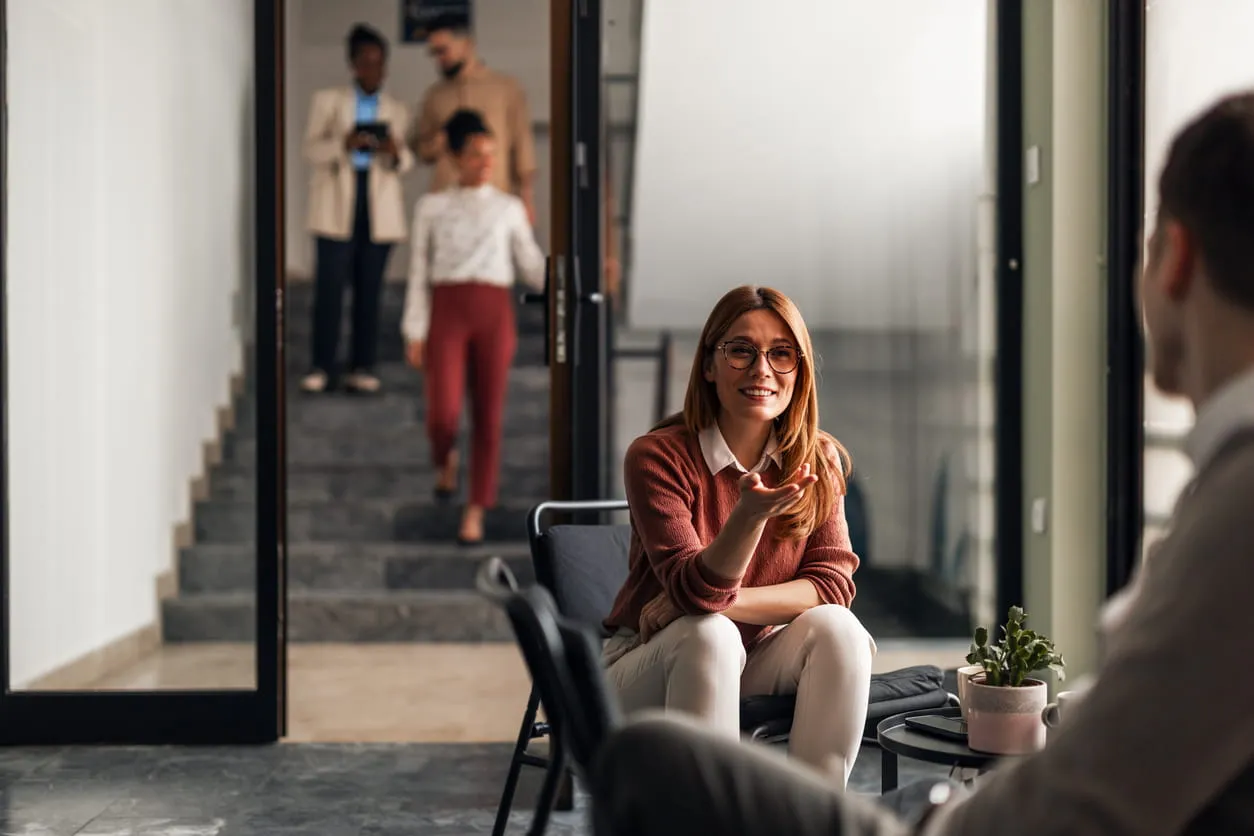 Woman with glasses and a rust-colored sweater talking to a man in an office.