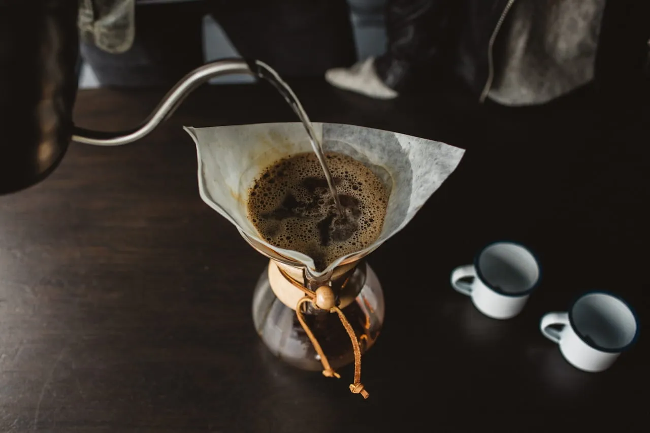 Water being poured into a coffee maker with two white ceramic mugs.