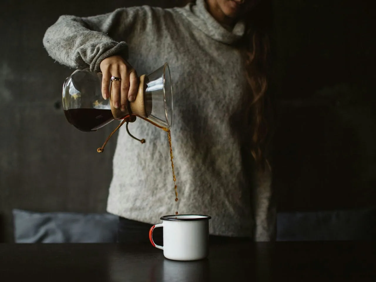 Person in a gray sweater pouring coffee from a glass carafe into a white mug.
