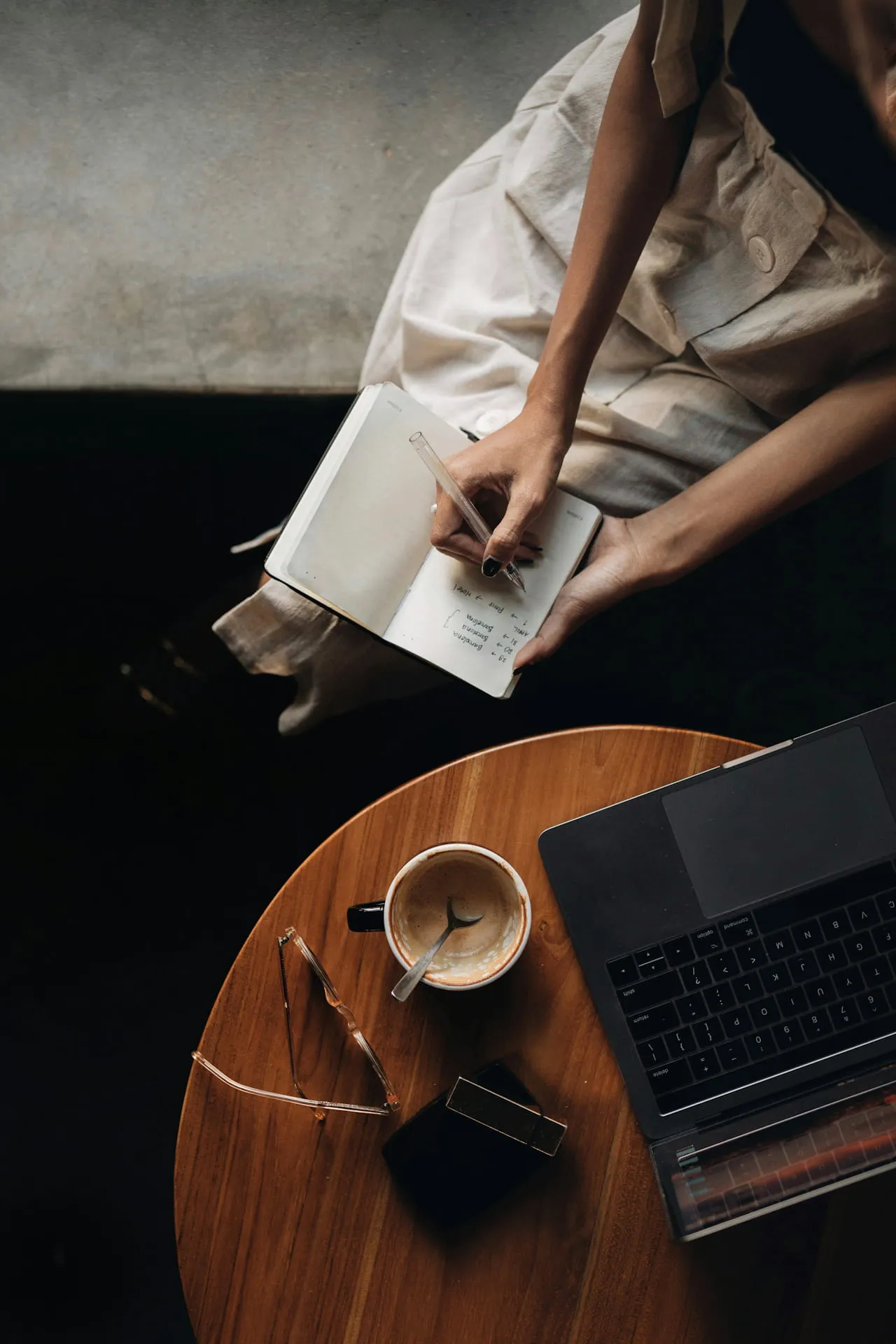 Person writing in a notebook while sitting next to a round wooden table with a cup, sunglasses, and a laptop.