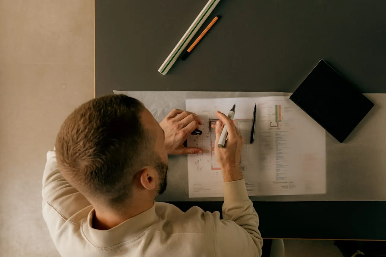 Man with short hair and beard drawing on architectural plans at a dark desk with markers and a black notebook.