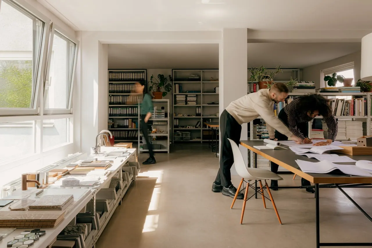 Two people leaning over a table with papers in a bright office, while another person walks in the background near shelves.