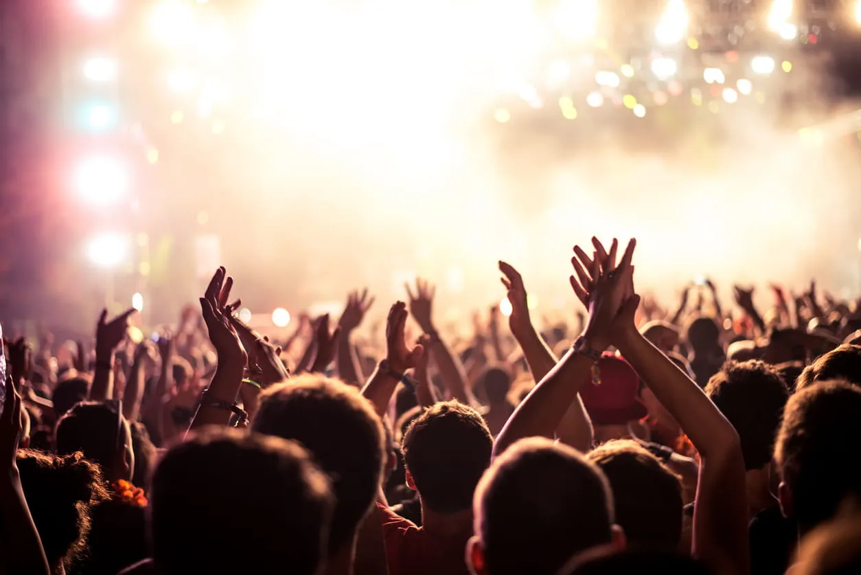 Crowd of people raising hands and clapping at a brightly lit concert or event.